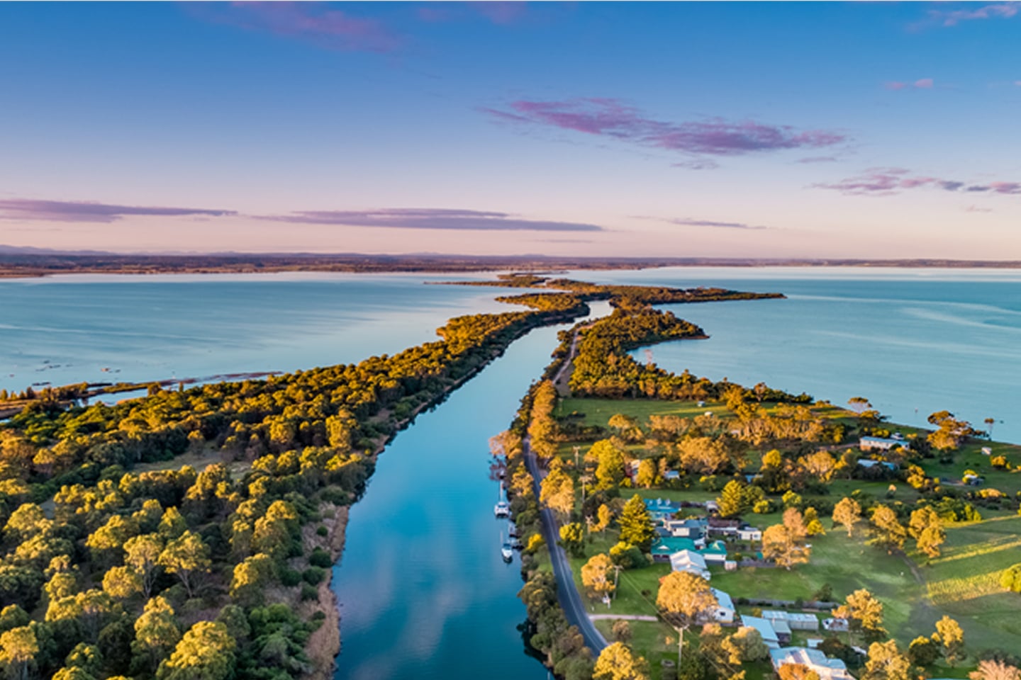 Aerial view of a narrow strip of land surrounded by calm blue water, with trees, houses, and a road.
